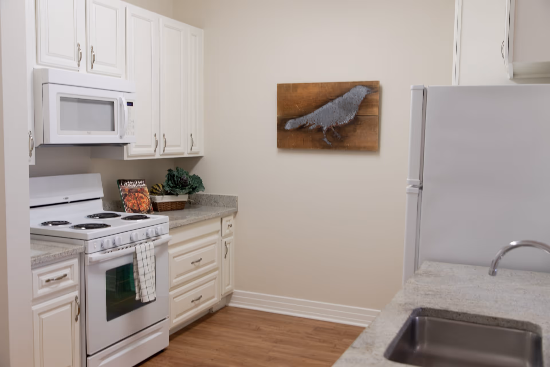 A clean kitchen with white cabinets, a white electric stove with oven, a white microwave above the stove, a white refrigerator, and a countertop with a sink. There is a decorative wooden wall art featuring a bird on the wall. The floor is wooden.