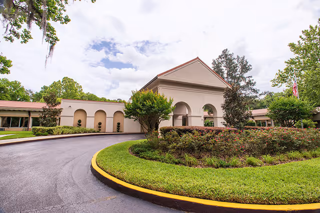 Exterior view of a single-story building with a curved driveway and landscaped greenery including bushes and trees under a partly cloudy sky.