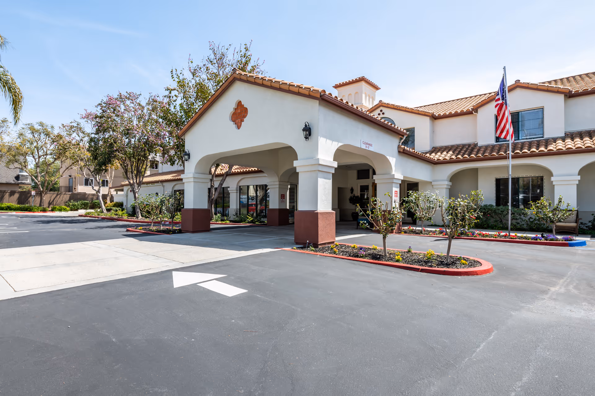 Front entrance of a Mediterranean-style senior living building with a covered porte-cochere, landscaped planters, and an American flag.