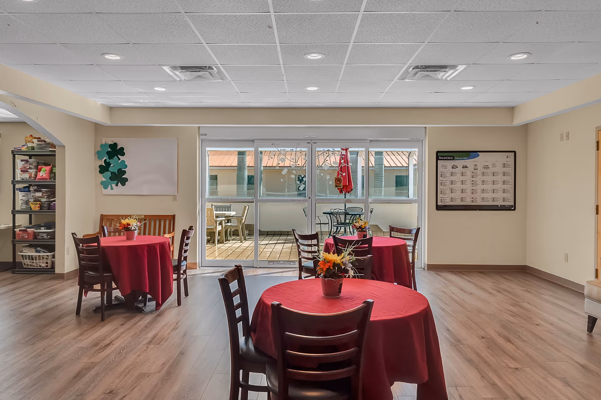 A bright dining area with three round tables covered in red tablecloths, each with a small flower arrangement. Wooden chairs surround the tables. Large sliding glass doors lead to an outdoor patio with additional seating. The room has light-colored walls, wood flooring, and a ceiling with recessed lighting. A shelving unit with various items is visible on the left, and a bulletin board is mounted on the right wall.