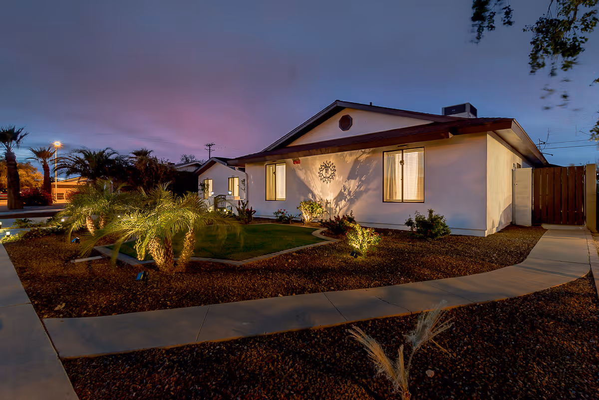 Exterior view of a single-story building at dusk with illuminated windows and landscaped garden featuring palm trees and shrubs, with a paved walkway in front.