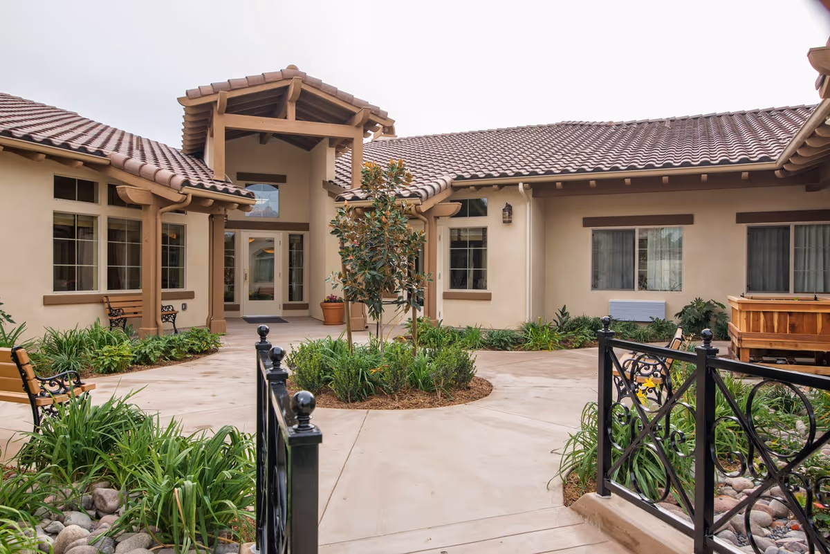 Outdoor courtyard area of a senior living facility with a central circular garden bed containing a small tree and plants. The courtyard is surrounded by beige buildings with tiled roofs and multiple windows. There are benches along the walkway and decorative black metal railings in the foreground.