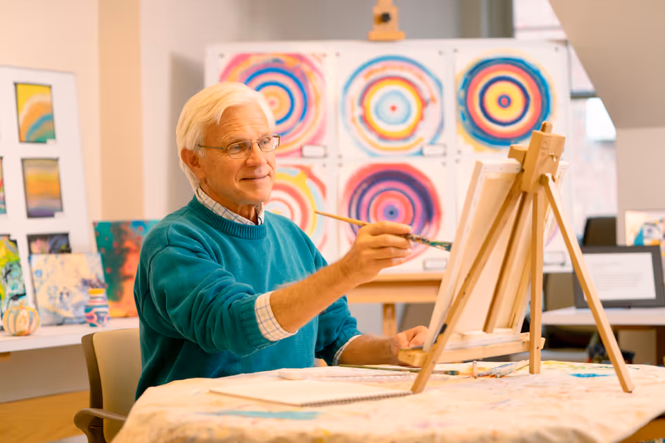 An elderly man with white hair and glasses is painting on a canvas set on an easel in a bright art studio. Behind him, colorful circular abstract paintings are displayed on the wall, and various other artworks are visible on a table. The man is wearing a teal sweater over a checkered shirt and appears focused and content while painting.