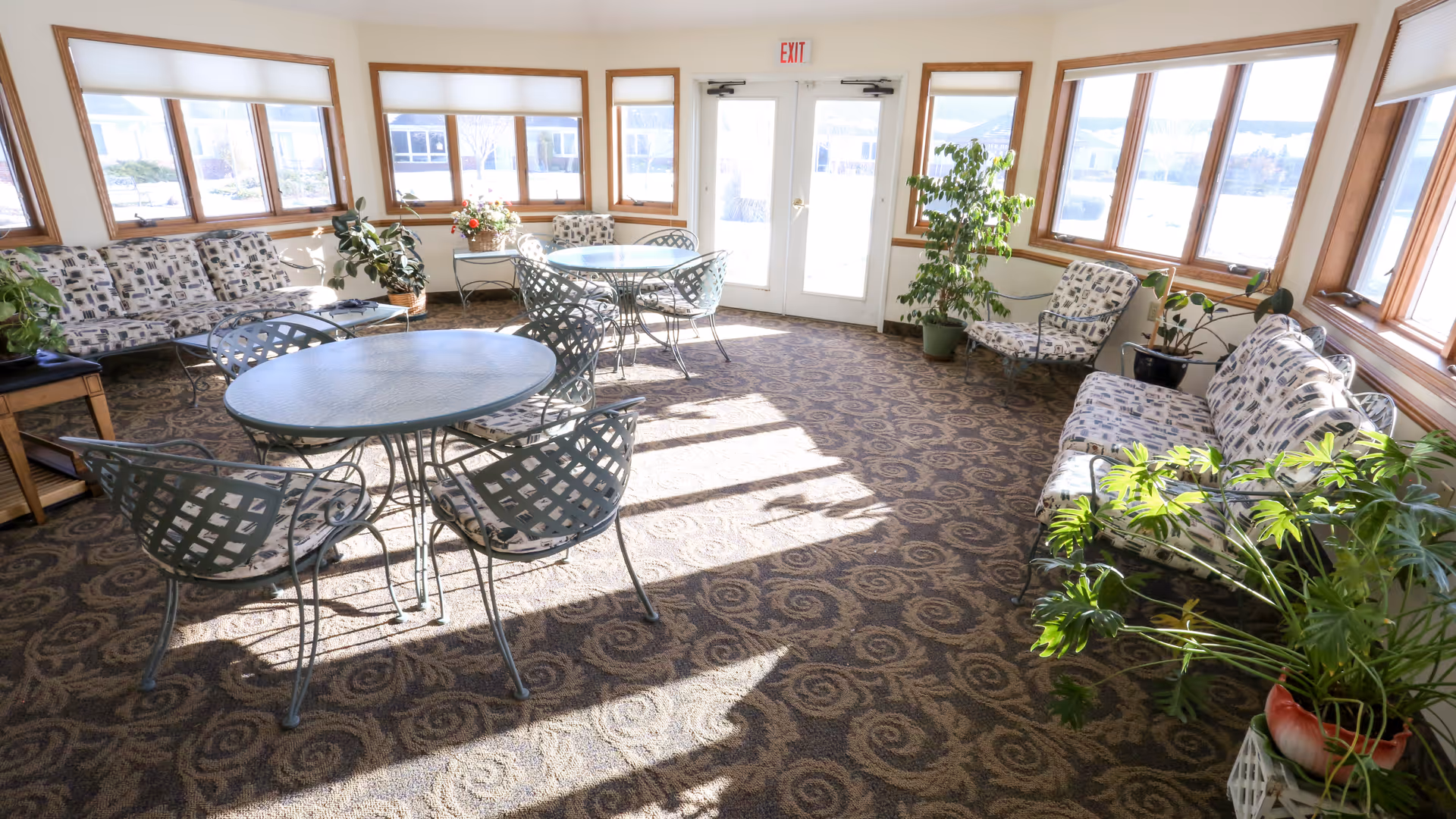 Bright sunlit common room with round metal tables, lattice chairs and cushioned sofas along windowed walls.