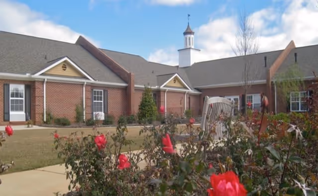 Exterior view of a single-story brick building with multiple windows and doors, a small cupola on the roof, and a garden with red flowers and a bench in the foreground under a partly cloudy sky.