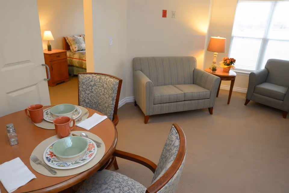 A cozy living area with a small round dining table set for two with plates, bowls, mugs, and utensils. The room features a striped loveseat, a gray armchair, a side table with a lamp and a flower pot, and a window letting in natural light. In the background, a bedroom with a bed and nightstand with a lamp is partially visible through an open door.