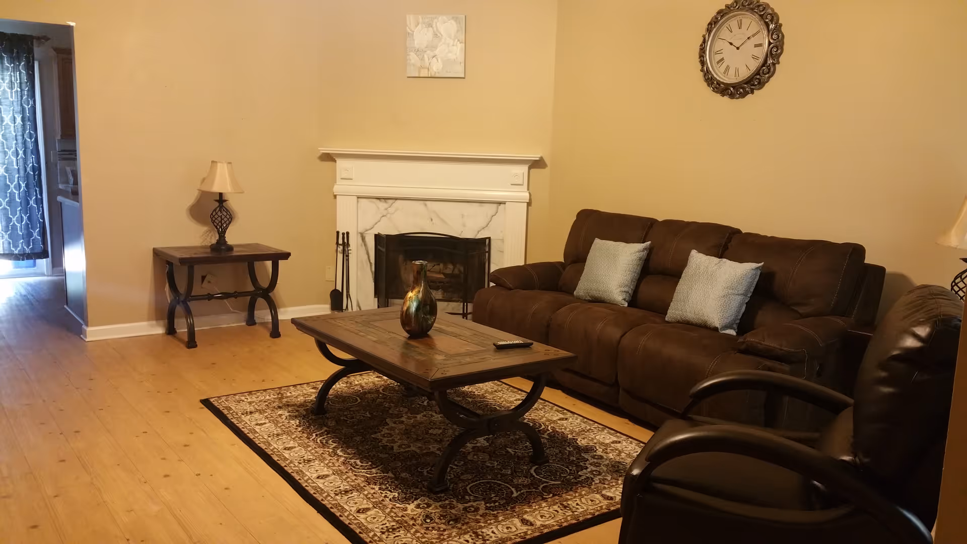 A cozy living room with a brown leather sofa adorned with two light blue pillows, a matching brown leather armchair, a wooden coffee table with a decorative vase, and a patterned area rug. There is a white fireplace with a marble surround, a wall clock above it, and a side table with a lamp. The room has light-colored walls and wooden flooring.
