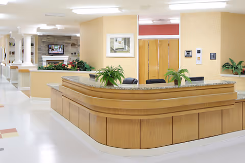 Reception area in a senior living facility with a wooden front desk, potted plants on the counter, beige walls, and a hallway leading to other rooms. A television is visible in the background near a fireplace.