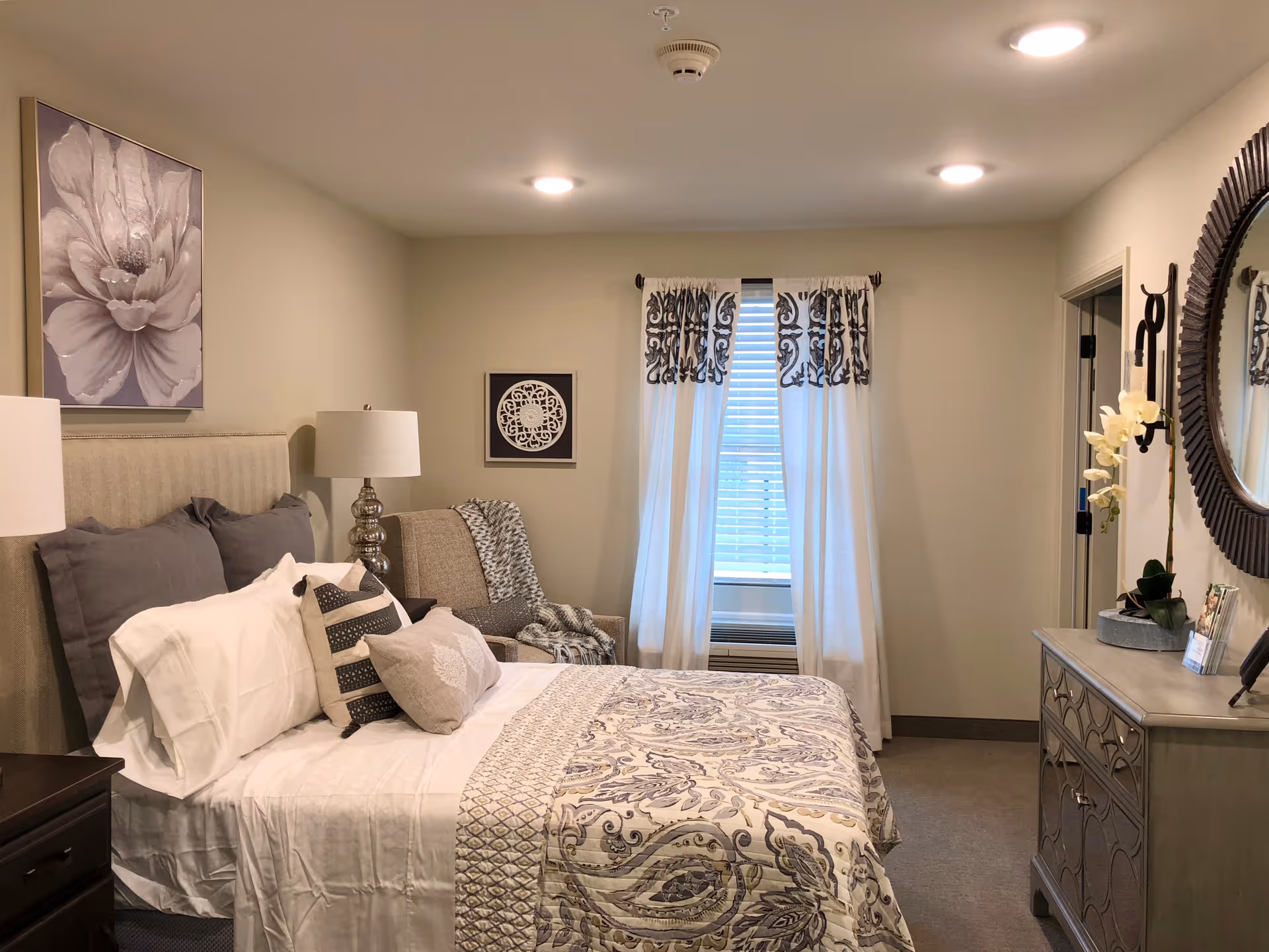 A cozy bedroom with a neatly made bed featuring white and patterned bedding, multiple pillows, and a beige upholstered headboard. There is a bedside table with a lamp, a comfortable armchair with a throw blanket, a window with white curtains and patterned valances, and a dresser with a round mirror and decorative items including a potted orchid.