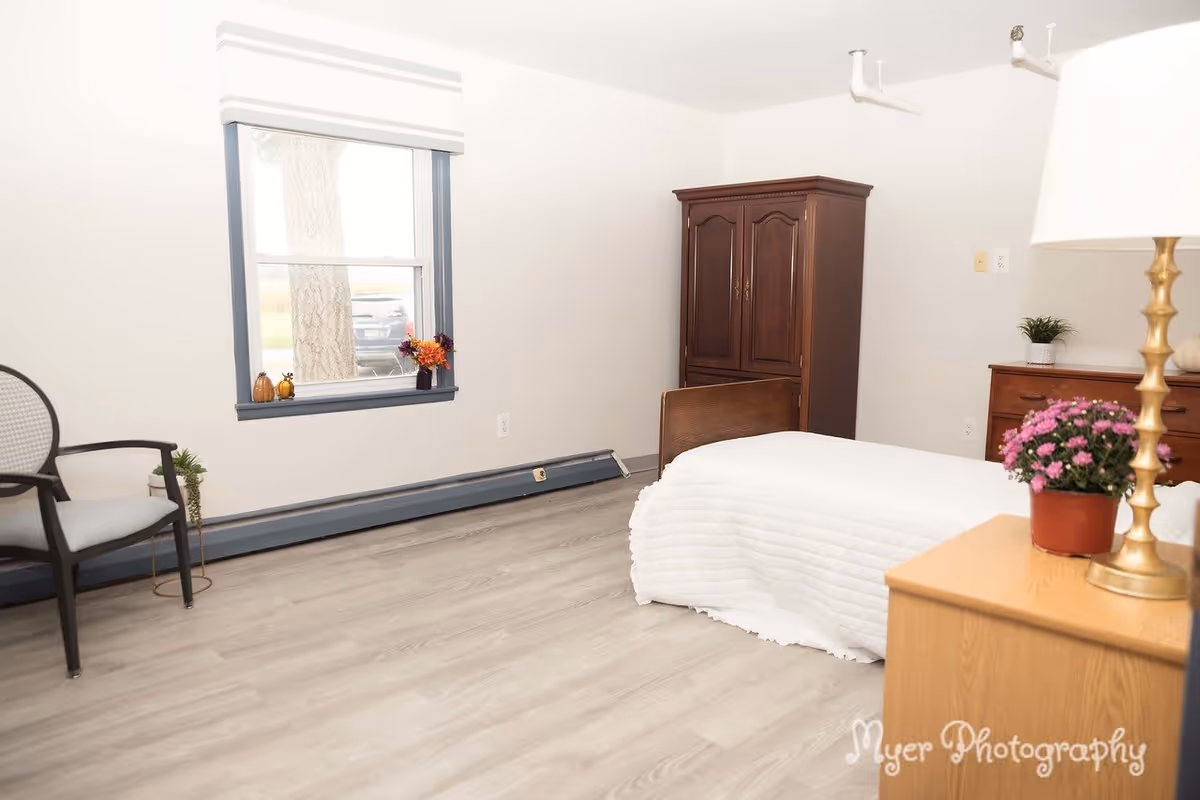 A simple bedroom with a single bed covered in a white quilt, a wooden armoire, a wooden dresser with a small plant, a chair, and a window with a white blind. There is a potted plant with pink flowers on a wooden nightstand beside the bed.