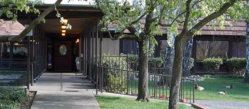 Entrance to Eskaton Care Center Greenhaven, with a covered walkway, railing, and surrounding trees.
