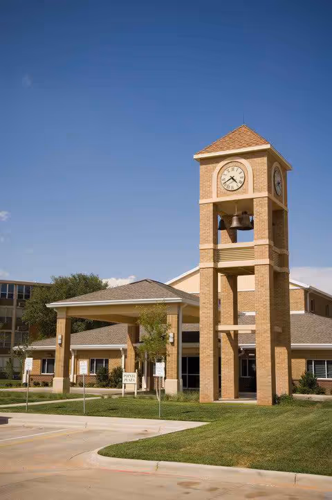 Exterior view of a senior living facility named Carillon featuring a tall brick clock tower with bells, a covered entrance with columns, and a well-maintained lawn under a clear blue sky.