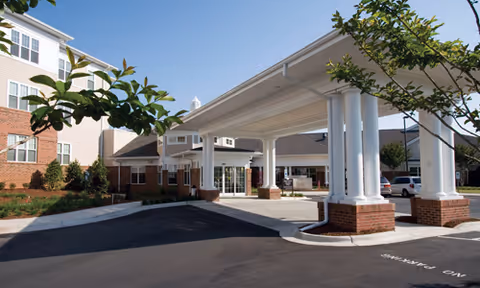Covered entrance (porte-cochère) of Croasdaile Village with white columns, brick bases, and the adjoining multi-story building under a blue sky.