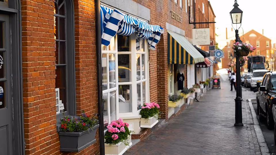 A charming street scene with brick buildings featuring colorful striped awnings and flower boxes filled with pink and purple flowers. The sidewalk is lined with vintage-style black street lamps adorned with hanging flower baskets. Cars are parked along the street, and a few people are visible in the distance.