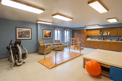 A rehabilitation room with exercise equipment including a stationary bike, parallel bars for walking practice, a padded therapy table, and an orange exercise ball. The room has blue walls, wooden cabinets along one side, and framed artwork on the walls. There are large fluorescent ceiling lights and windows with valances.