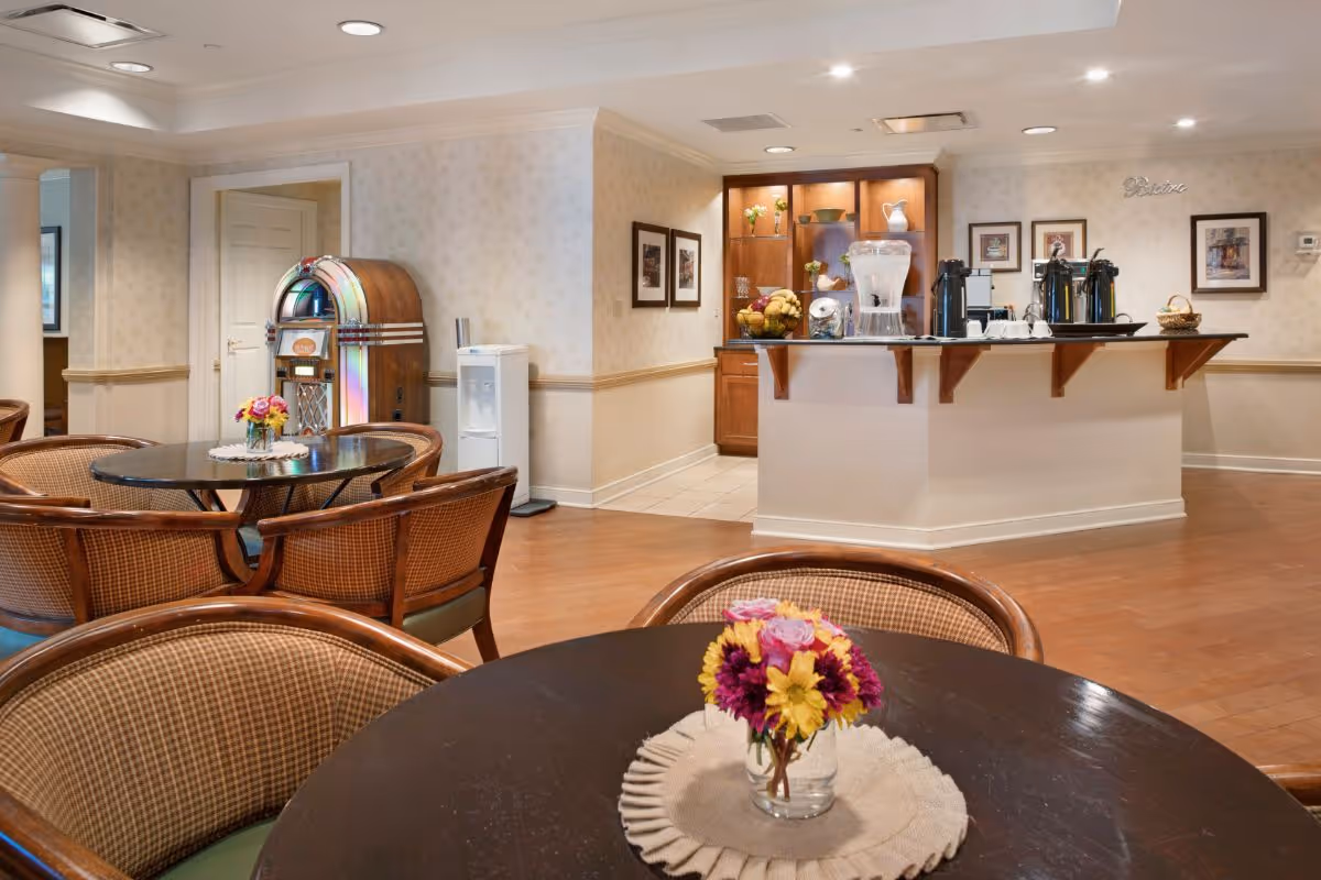 Interior view of a common area in a senior living facility featuring round tables with floral centerpieces, wicker chairs, a vintage jukebox, a water dispenser, and a counter with coffee urns and a water pitcher. The walls are decorated with framed pictures and soft lighting.