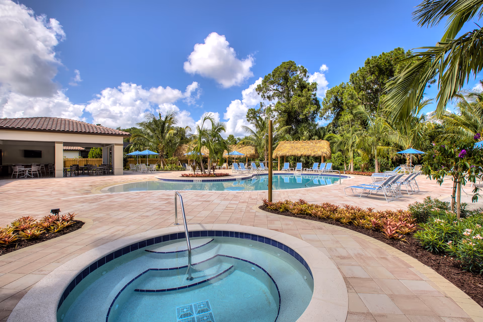 Outdoor pool area at The Arlington of Naples featuring a circular hot tub in the foreground, a larger swimming pool with lounge chairs and umbrellas, tropical palm trees, and a covered seating area under a clear blue sky with scattered clouds.