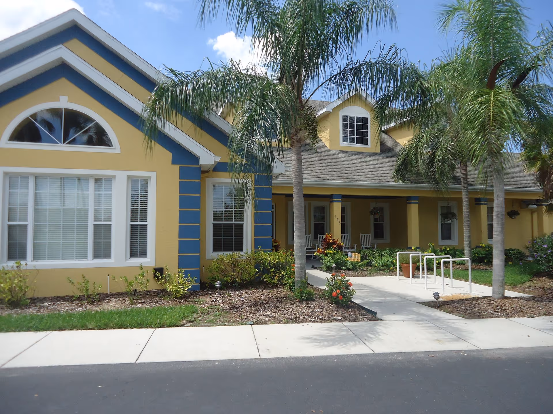 Exterior view of a yellow and blue senior living facility building with a covered porch, palm trees, and landscaped garden beds along a sidewalk.
