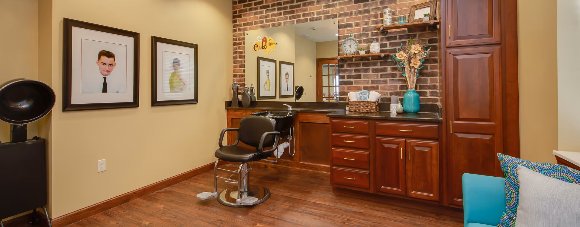 Interior of a hair salon area with a black salon chair in front of a sink, wooden cabinets with drawers, a large mirror on a brick wall, decorative shelves with plants and a clock, framed vintage portraits on the wall, and a teal chair with patterned pillows.