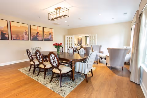 A bright and spacious dining area in a retirement community featuring a large wooden dining table with eight cushioned chairs, a vase of red flowers as centerpiece, a patterned rug underneath, and a seating area with four upholstered armchairs near large windows with curtains. The walls are decorated with three framed pictures, and there is a modern rectangular ceiling light fixture.