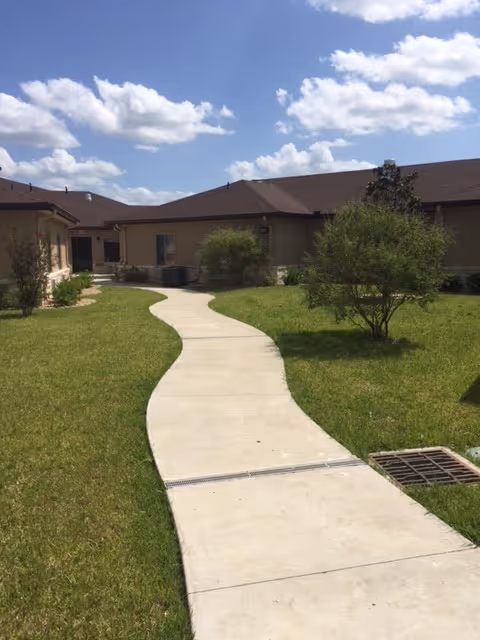 A winding concrete pathway through a grassy courtyard with small bushes and trees, surrounded by single-story beige buildings under a partly cloudy blue sky.