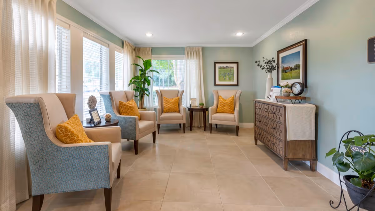 Bright communal seating area with upholstered armchairs, yellow accent pillows, side tables, plants, and a decorative console against a pale green wall.