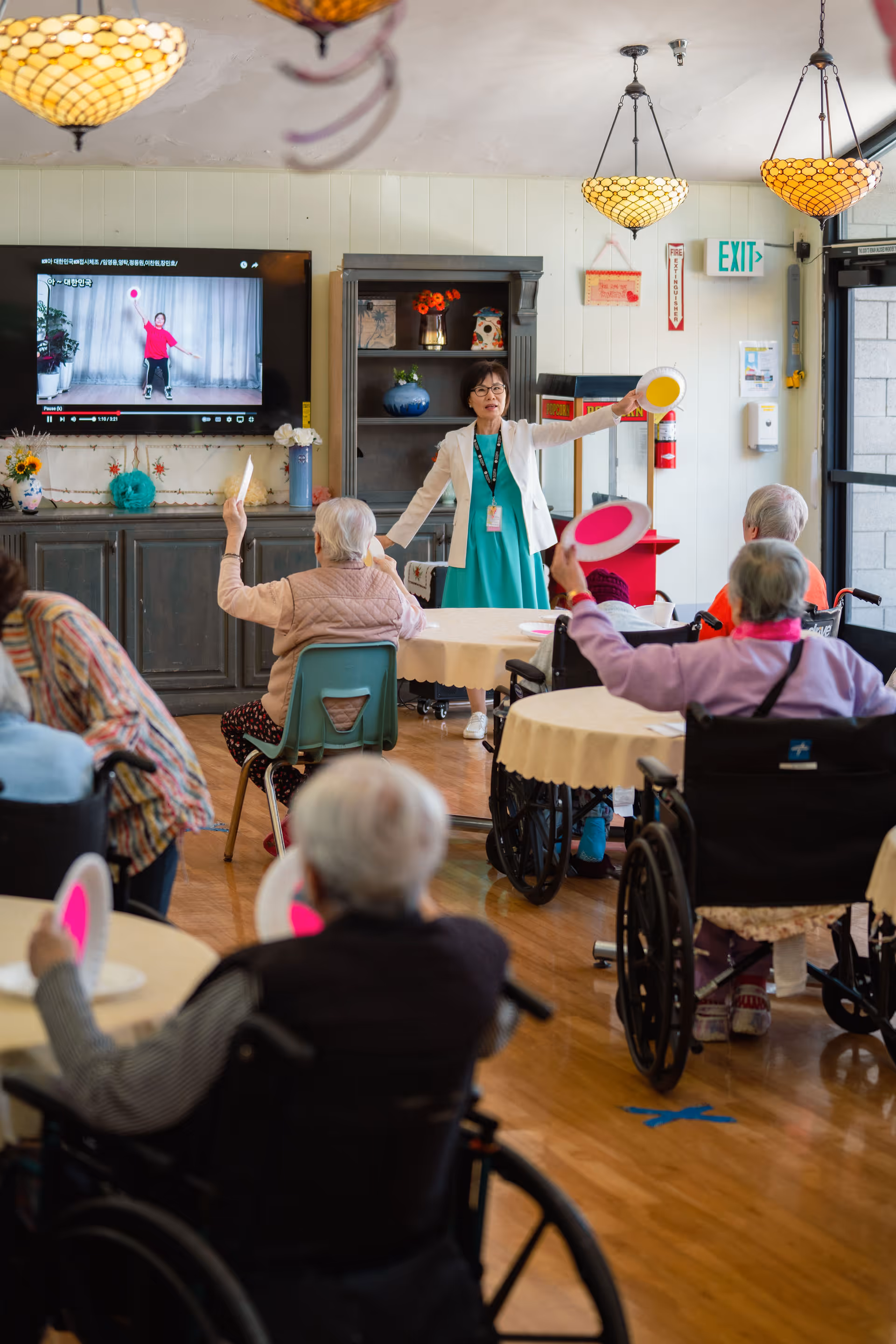A group of elderly individuals, some in wheelchairs, participate in a seated exercise activity led by a woman standing in front of them holding colorful paddles. A large TV screen on the wall shows a person demonstrating an exercise with a red ball. The room has round tables with white tablecloths, warm lighting fixtures, and a cabinet with decorative items.
