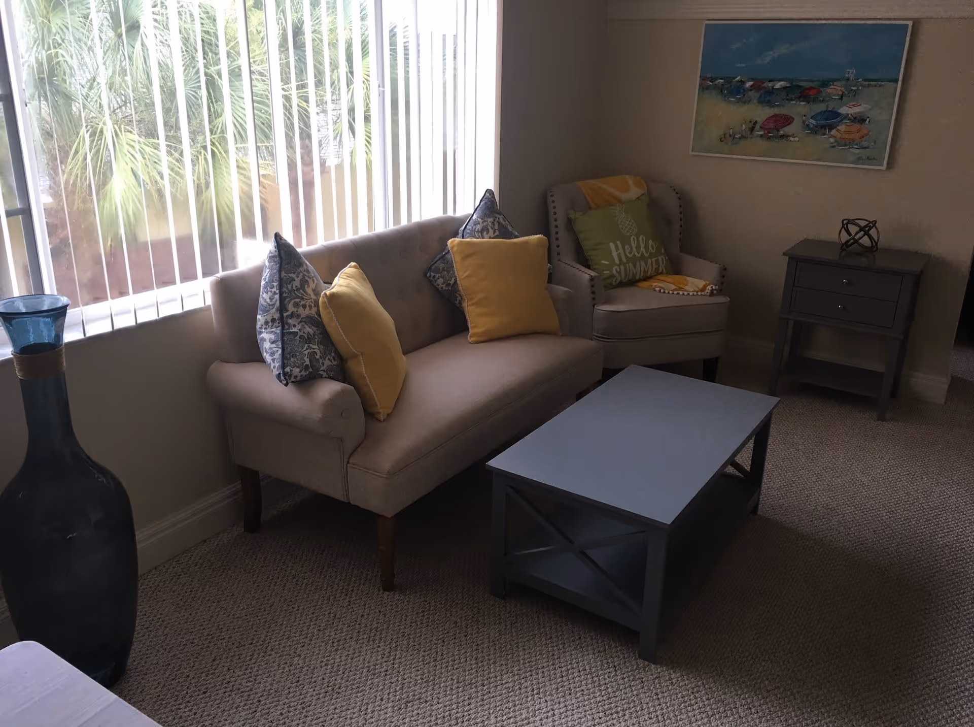 Small living room with a sofa, armchair, coffee table, side table and a large window with vertical blinds.