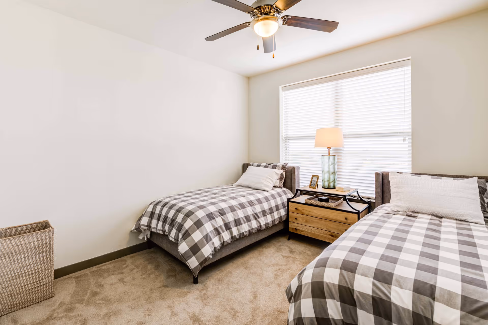 Two twin beds with gray plaid bedding in a bright, neutral bedroom with a nightstand, lamp, blinds, and a ceiling fan.