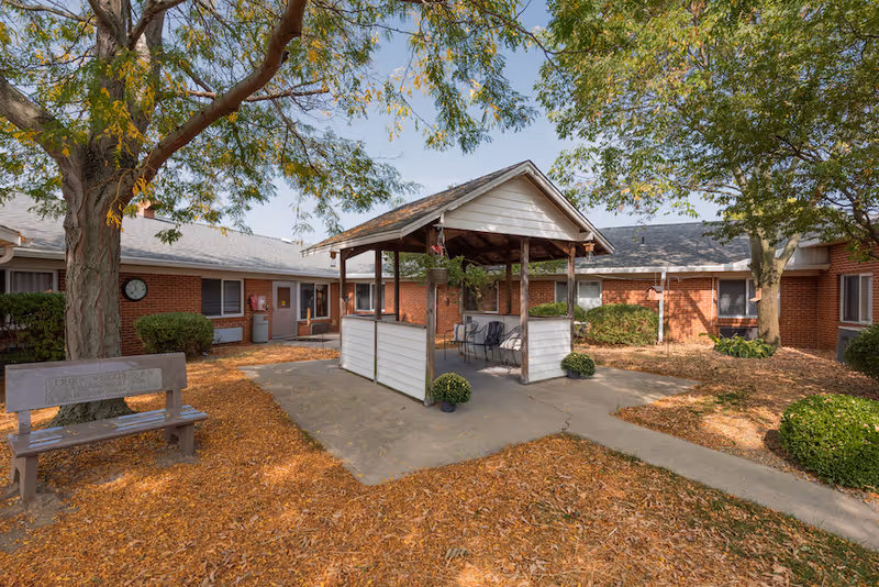 Courtyard of a single-story brick rehabilitation center with a covered gazebo seating area, benches, trees, and fallen leaves.