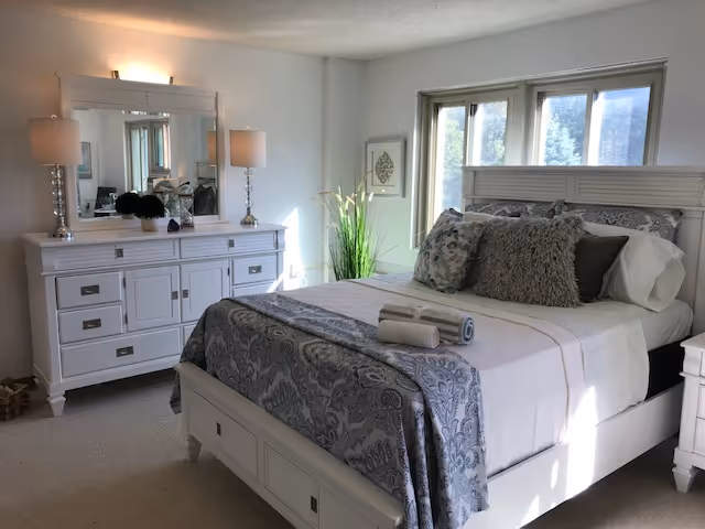 Sunlit bedroom featuring a white bed with patterned bedding and throw pillows, a matching dresser with mirror and lamps, and windows.