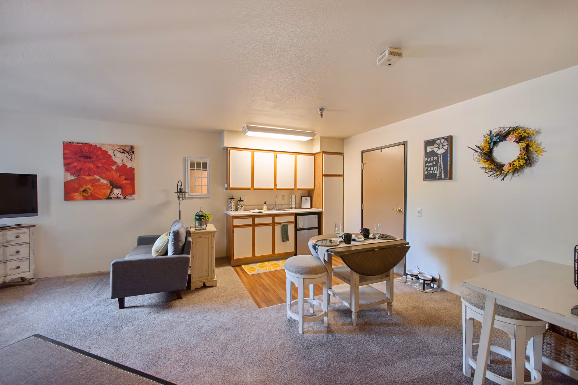 Interior view of a senior living apartment featuring a small kitchen area with white cabinets and wooden trim, a dining table set for two with stools, a gray couch with pillows, a TV on a white dresser, and decorative wall art including a red flower painting and a wreath.