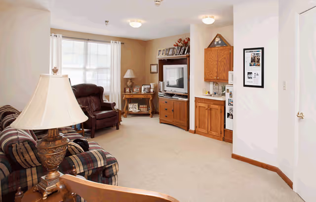 A cozy living room area with a plaid sofa, a brown leather armchair, a wooden TV cabinet with a television, a small side table with a lamp, and a kitchenette with wooden cabinets and a small refrigerator. The room has beige walls, carpeted floor, and a window with white curtains letting in natural light.