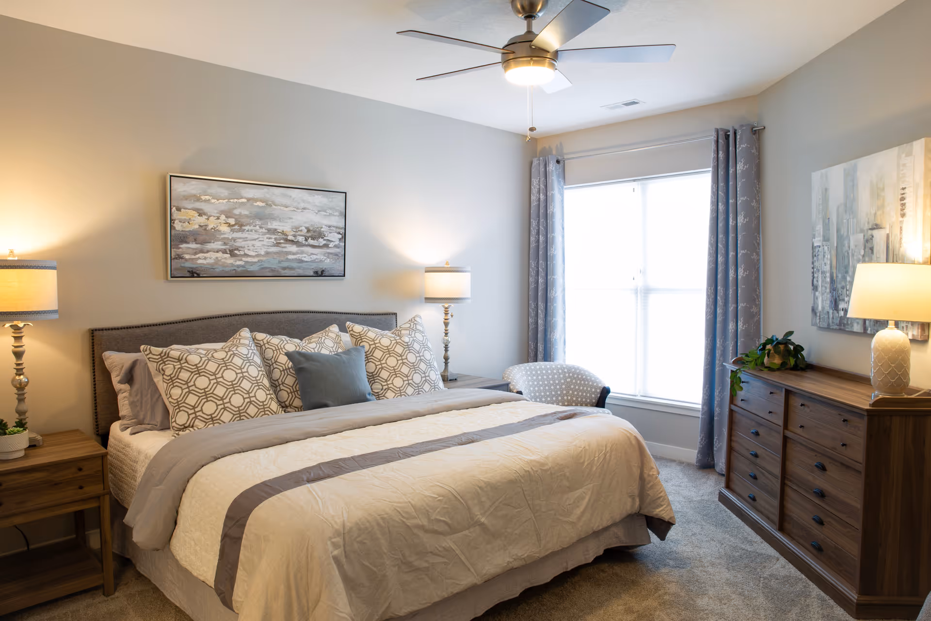 A cozy bedroom with a large bed featuring multiple patterned pillows and a beige comforter with a gray stripe. There are two wooden nightstands on either side of the bed, each with a lamp. A ceiling fan with a light is above the bed. A window with blue patterned curtains lets in natural light. A wooden dresser with a lamp and a plant is positioned against the wall, and two pieces of abstract artwork hang on the walls.