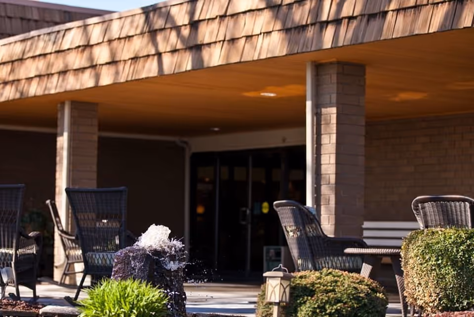 Covered outdoor seating area with wicker chairs, shrubs and a small fountain at the entrance of a retirement facility.