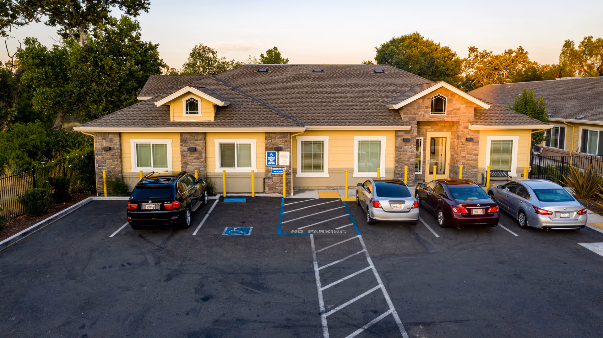 Front exterior view of a single-story senior living facility building with a yellow and stone facade, several windows, and a main entrance door. There are five cars parked in front of the building, including one in a handicapped parking space. The parking lot is marked with white and blue lines, and there are trees and greenery surrounding the building.