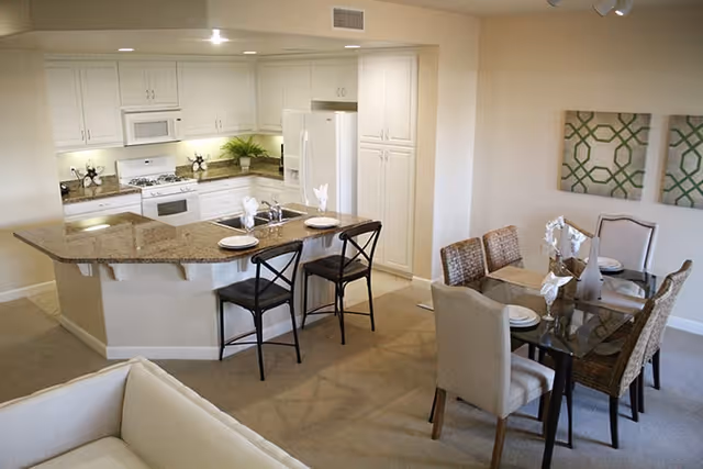 A bright and clean kitchen and dining area in a senior living facility. The kitchen features white cabinets, a white refrigerator, a white stove with a microwave above it, and a granite countertop with two black chairs at a breakfast bar. The dining area has a glass table with six chairs, set with plates and napkins. Two decorative green and white patterned wall hangings are visible on the wall behind the dining table.