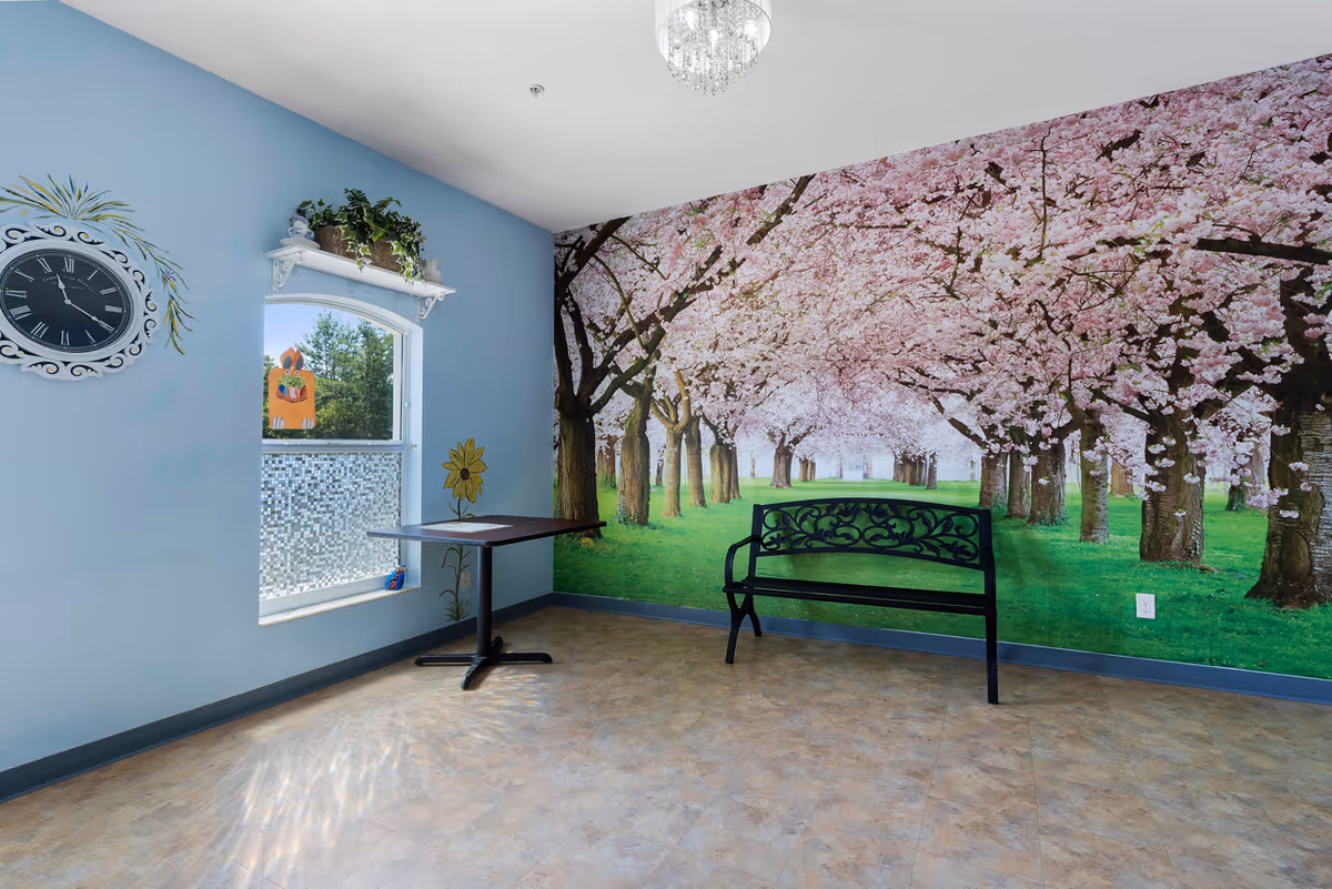 A small room with light blue walls featuring a large mural of cherry blossom trees in full bloom along one wall. There is a black decorative bench against the mural wall and a small table near a window with a white decorative shelf above it holding potted plants. A round wall clock with Roman numerals is mounted on the left wall, and a small chandelier hangs from the ceiling.