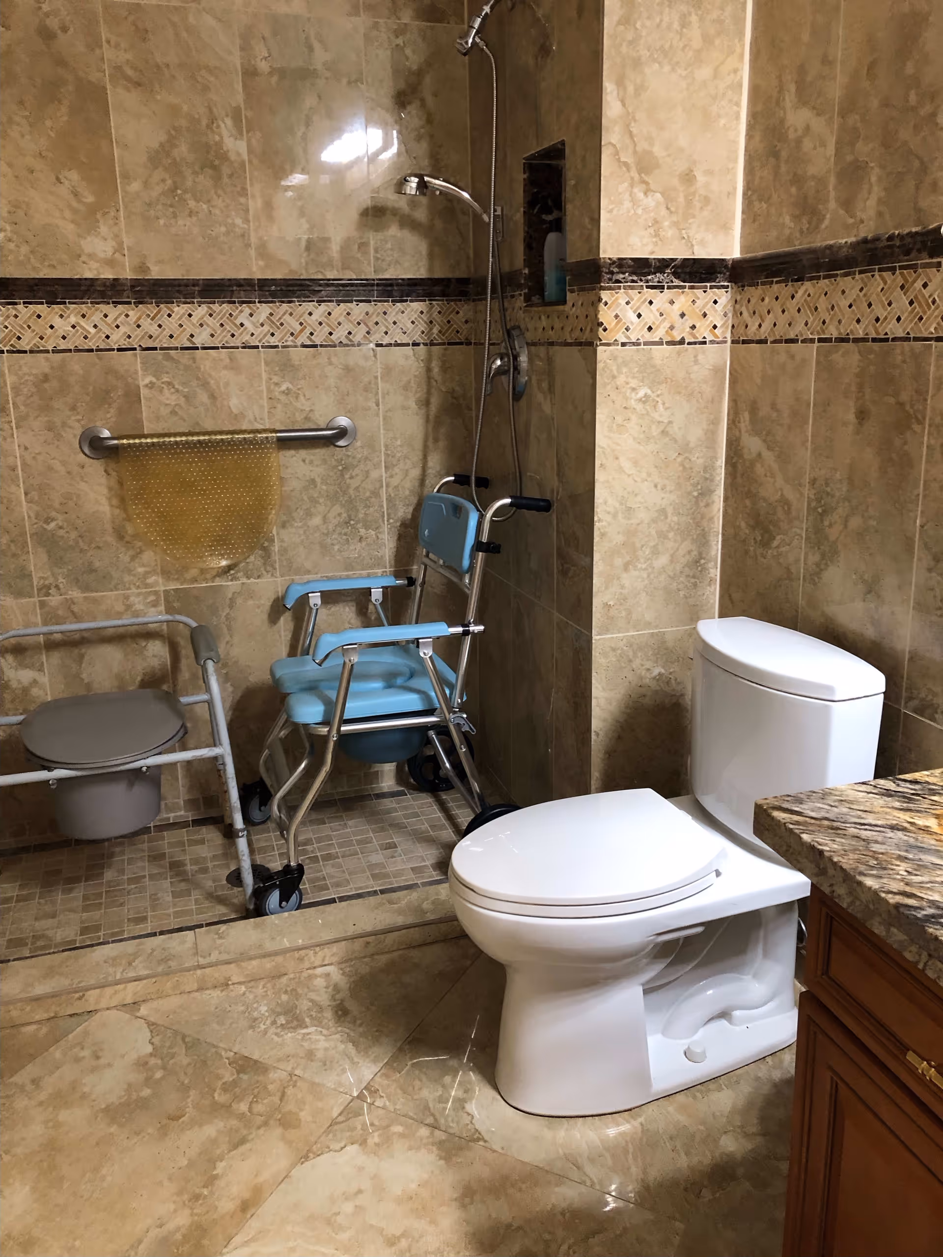 A bathroom with beige tiled walls and floor, featuring a white toilet, a granite countertop with wooden cabinetry, and a walk-in shower area equipped with a blue shower chair and a gray commode chair. There is a metal grab bar with a yellow textured mat hanging on it inside the shower.