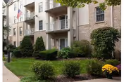 Exterior view of a multi-story residential building with balconies, surrounded by well-maintained shrubs, bushes, and a small tree. An American flag is visible near the entrance, and a sidewalk runs along the front of the building.