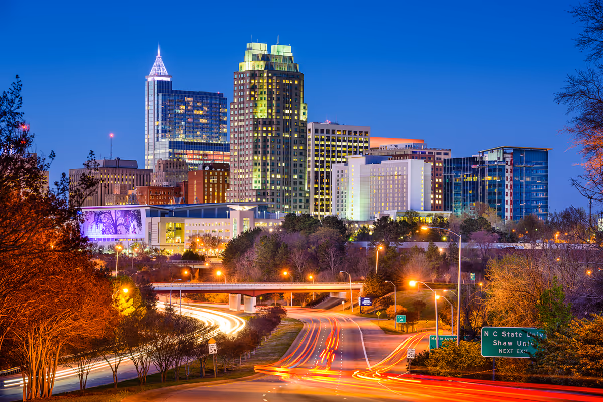 Evening cityscape of downtown Raleigh with illuminated skyscrapers and light trails from vehicles on a highway in the foreground.