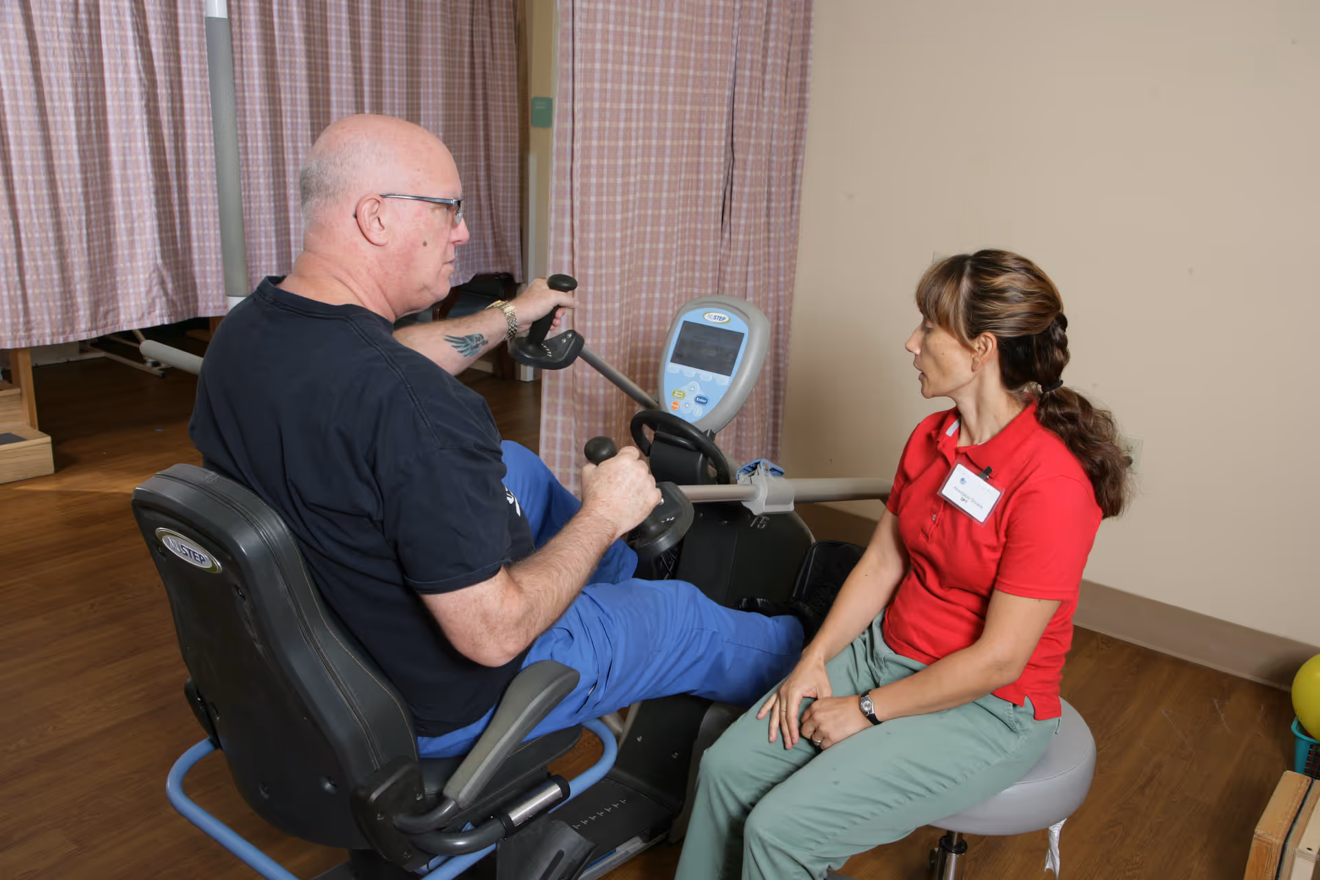 An elderly man using a seated exercise machine while a female caregiver or therapist in a red shirt and green pants sits nearby, observing and assisting him in a room with wood flooring and pink checkered curtains.