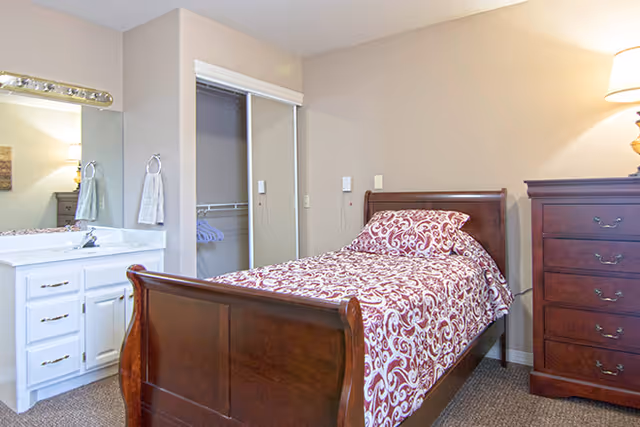 Single bedroom with a wooden sleigh bed dressed in red patterned bedding, a tall dresser, open closet and a vanity sink.
