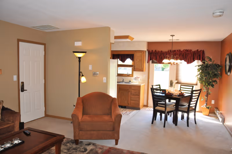 Open-concept interior showing a living area with an armchair and coffee table, a dining table, and a small kitchen in the background.