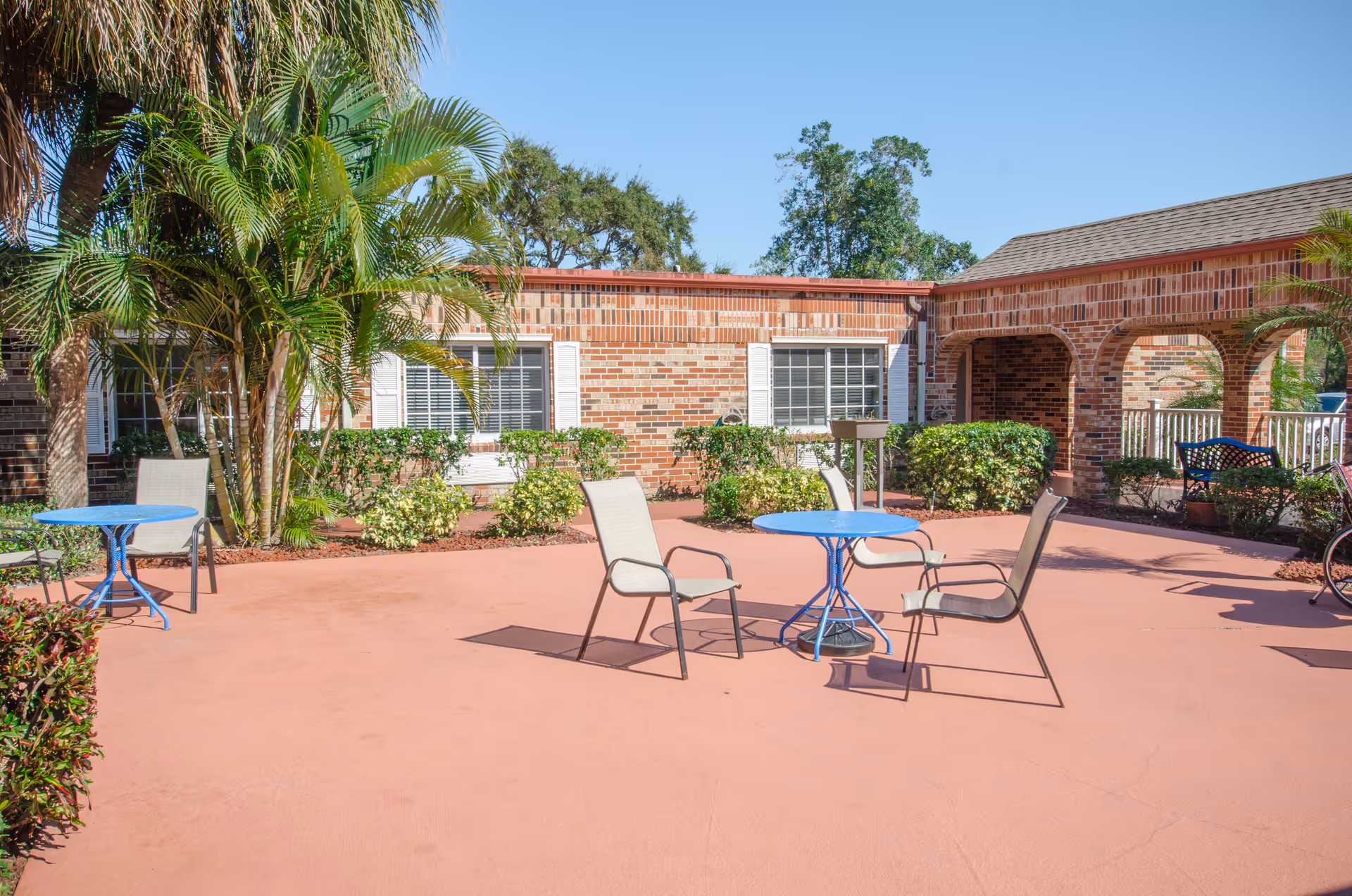 Outdoor patio area at Anchor Care and Rehabilitation Center with blue round tables and beige chairs on a reddish concrete floor. The patio is surrounded by brick buildings with white window shutters, green bushes, and palm trees under a clear blue sky.