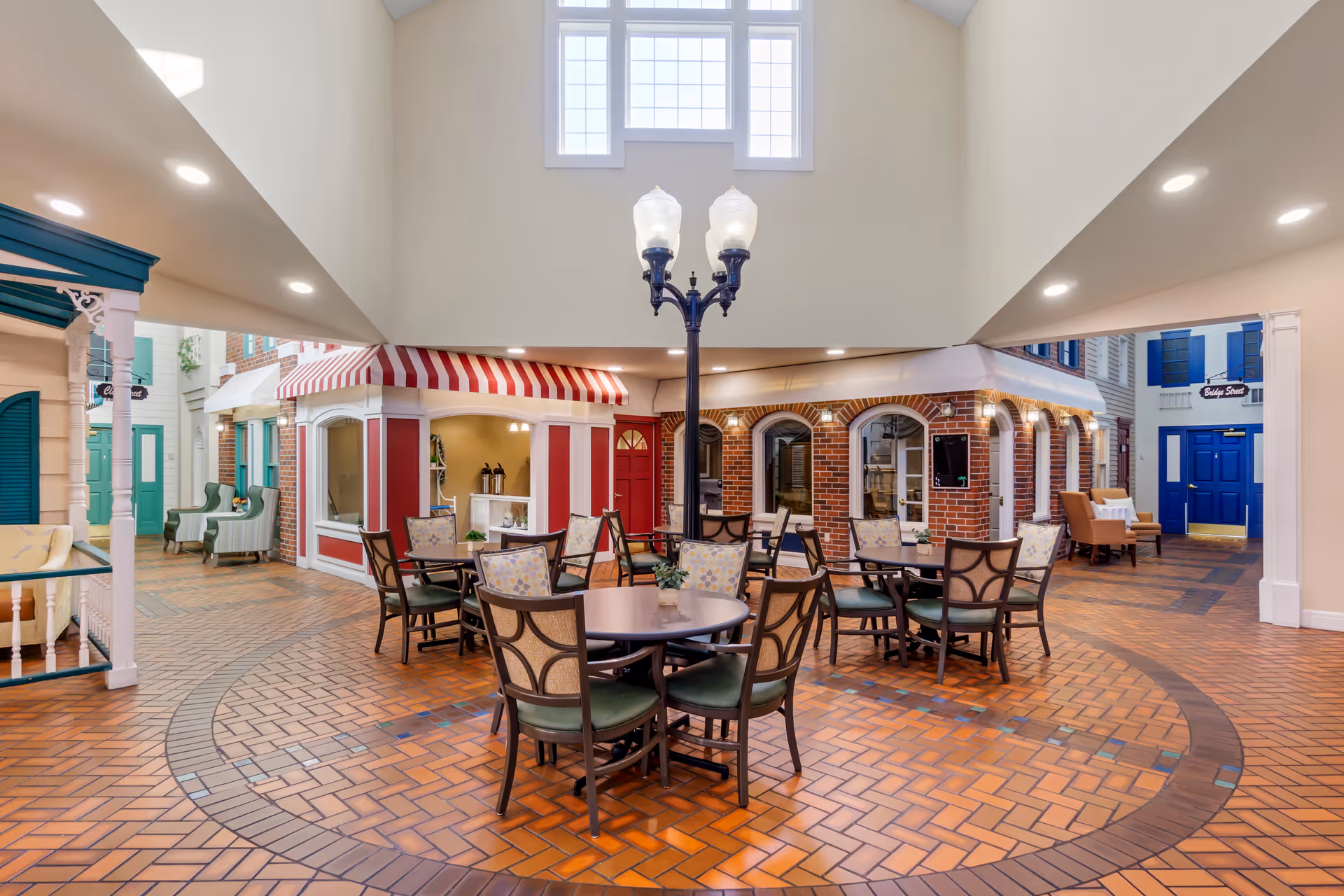 Indoor town-square style common area with round dining tables and chairs, storefront facades, and a central lamppost under a skylight.