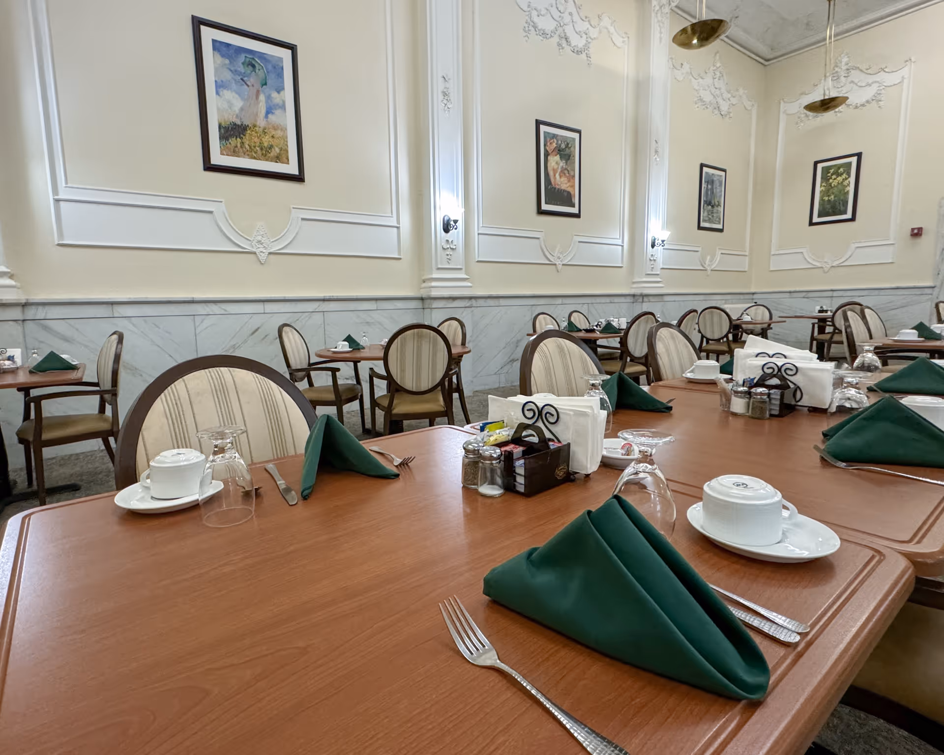A dining room with multiple wooden tables set with green folded napkins, white cups and saucers, upside-down glasses, silverware, and condiment holders. The room has high ceilings with ornate molding, cream-colored walls with framed paintings, and upholstered chairs with wooden frames.