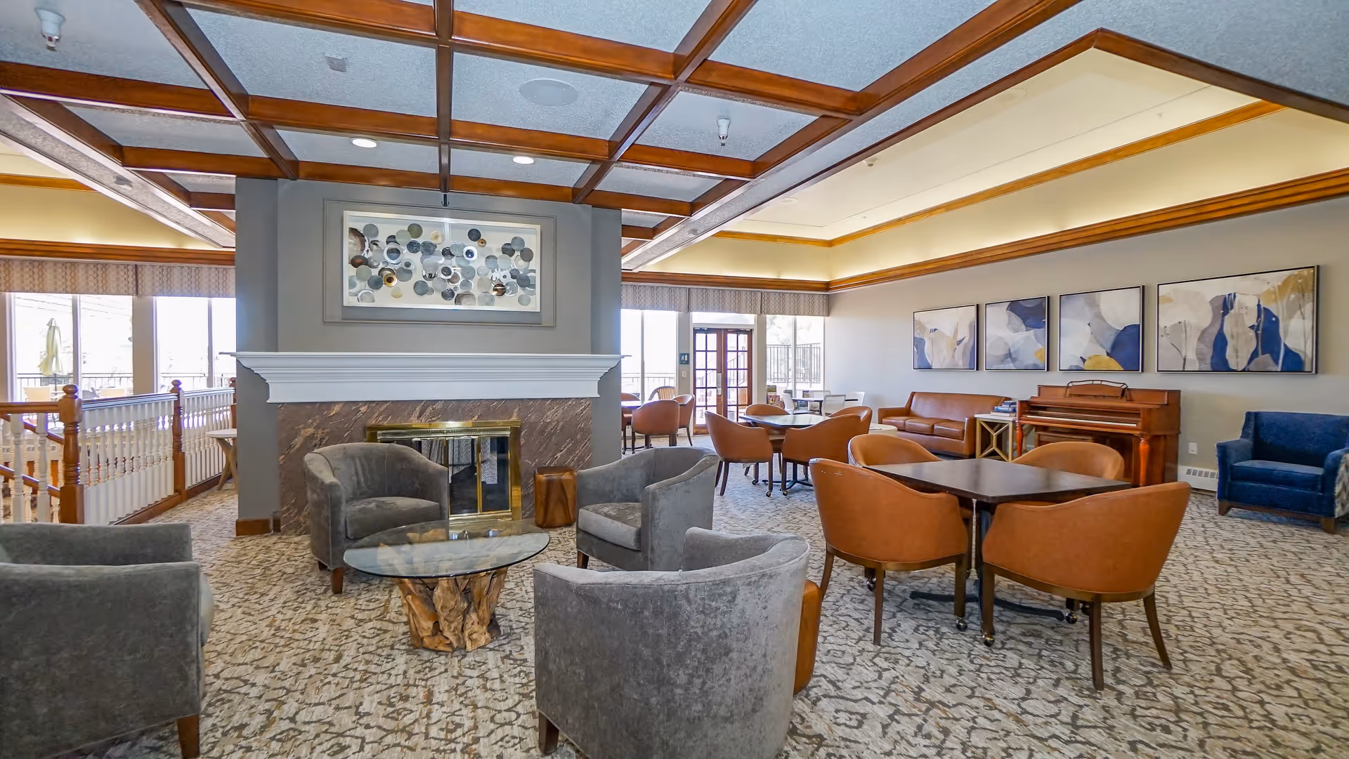 A spacious and well-lit common area with a fireplace in the center. The room features several seating arrangements including gray armchairs around a glass coffee table with a wooden base, brown chairs around dark wood tables, a brown leather sofa, and a blue armchair. There is a piano against the wall with abstract artwork hanging above it. Large windows and a glass door allow natural light to fill the room.