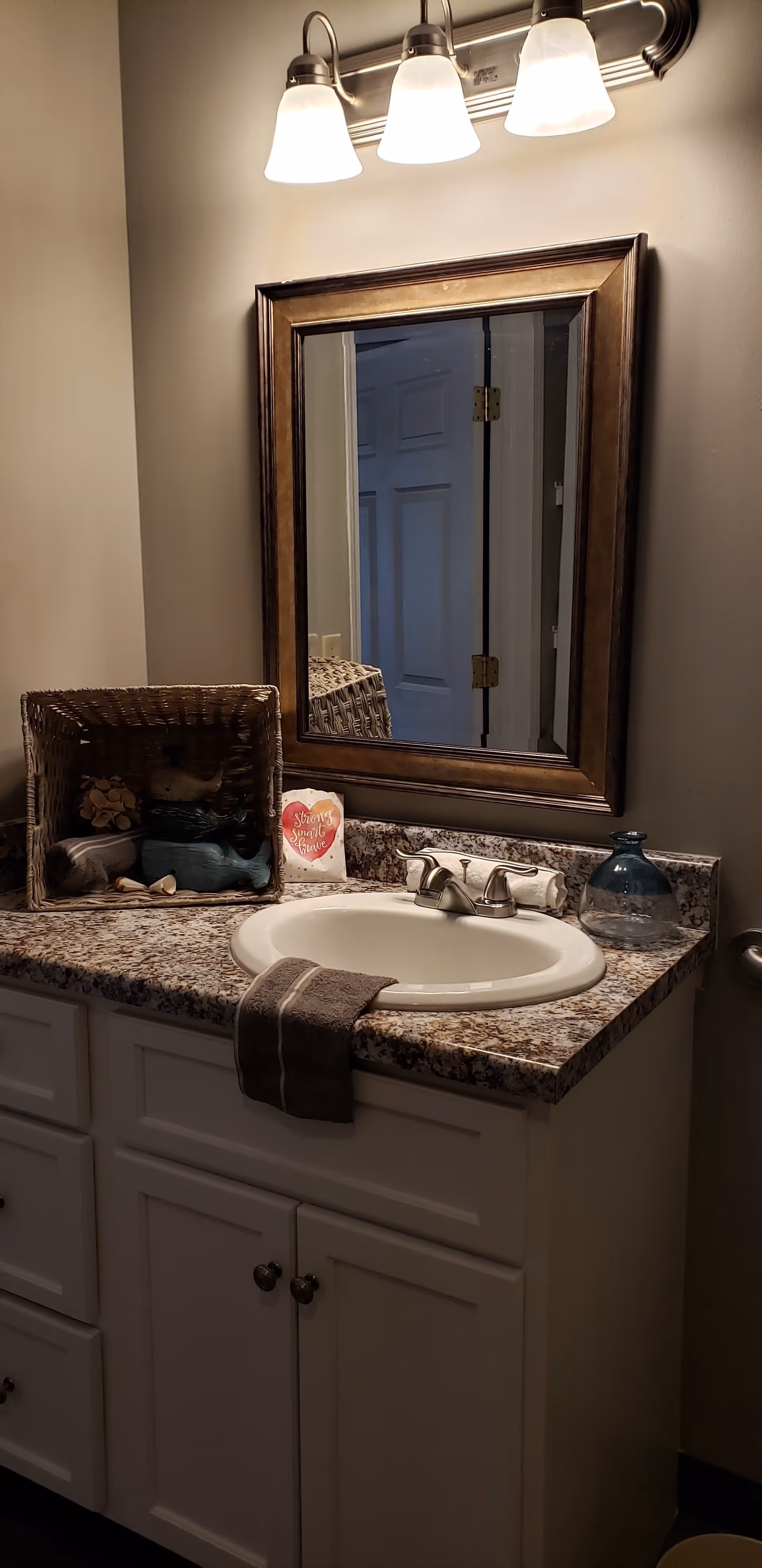 Bathroom vanity with a granite countertop, white sink, and silver faucet. Above the sink is a rectangular mirror with a brown frame and a three-light fixture with frosted glass shades. On the countertop, there is a wicker basket with decorative items, a small sign that reads 'Strong Smart Brave,' a rolled white towel, and a blue glass vase. A dark gray hand towel hangs over the edge of the countertop.