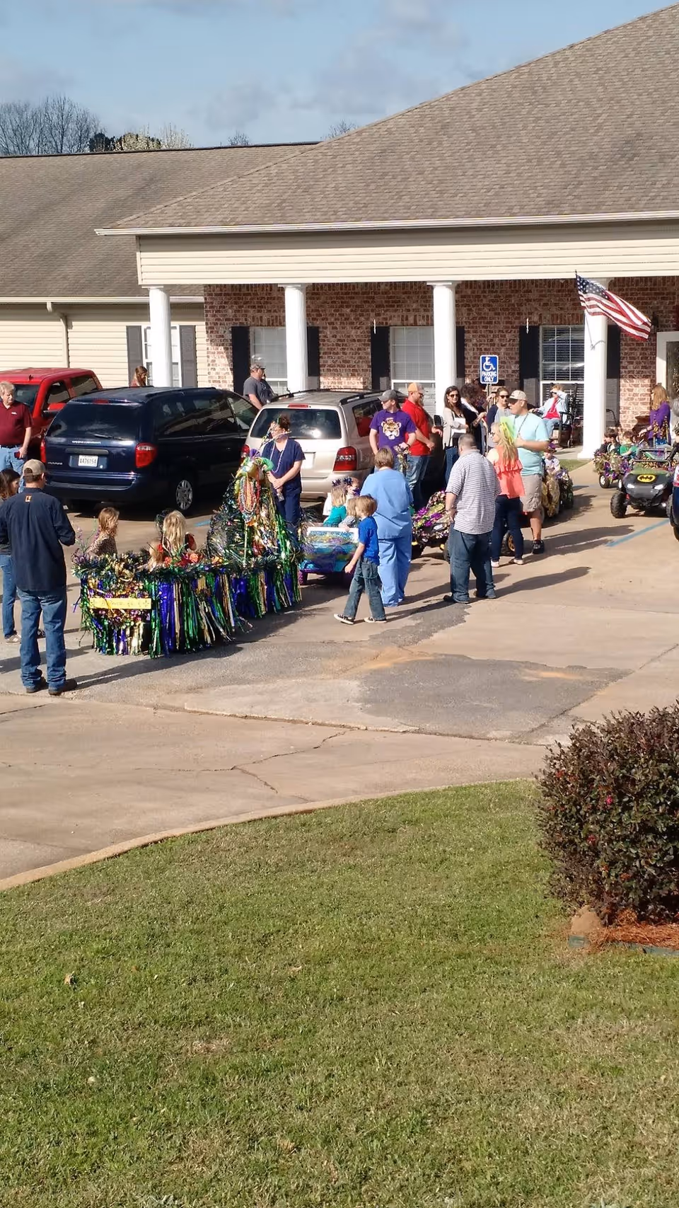 A group of people, including children and adults, gathered outside a brick building with white columns. Some children are riding in small decorated carts or wagons, and adults are standing nearby. There are several parked vehicles, an American flag, and a handicap parking sign visible near the building entrance.